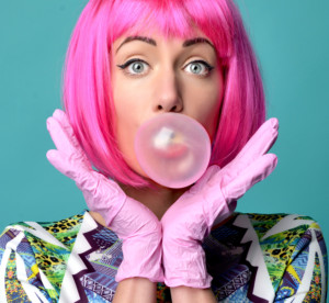 Close up funny fashion portrait of cheerful woman inflating the bubble gumin hot pink party wig on a mint background.