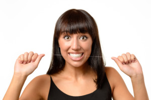 Excited attractive tanned young woman with a wide eyed look of amazement holding a string of dental floss in her clenched teeth, head and shoulders facing the camera